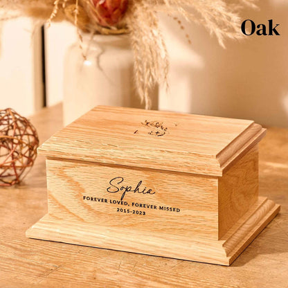 An personalised children's cremation urn sits on a wooden table. The urn is rectangular and is engraved on the front face with the words "Sophie - Forever Loved, Forever Missed 2015-2023". On the top face is a large capital letter S which has a floral design. It is an urn for human ashes.