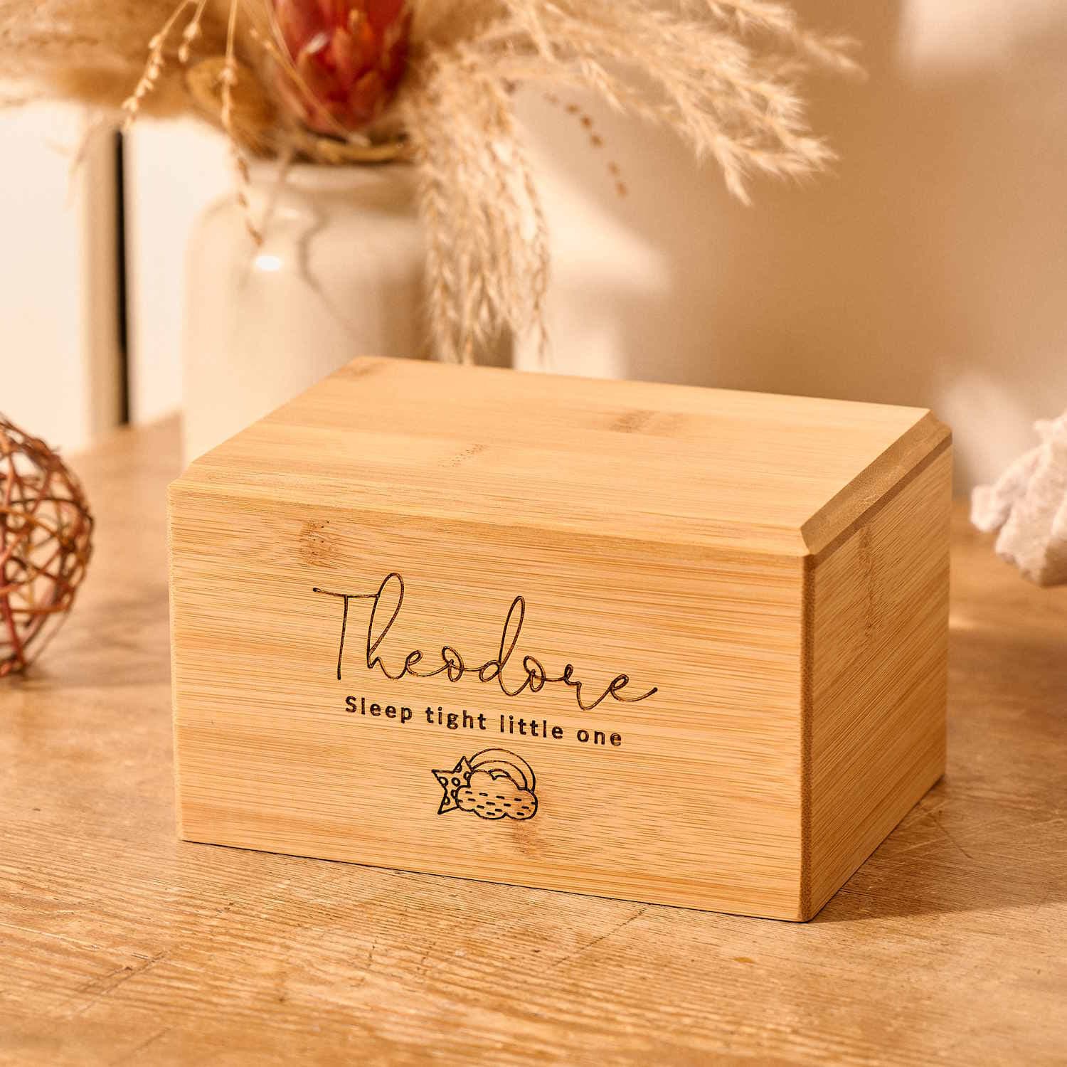 A small cremation urn for a stillborn baby or a miscarriage sits on a table. It is engraved with a cloud and rainbow motif and the name "theodore". A message reads "sleep tight little one". It is a personalised cremation urn.